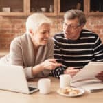Retirement Financial Planning Concept. Happy Senior Couple Discussing Family Budget Together, Sitting In Kitchen With Laptop And Papers